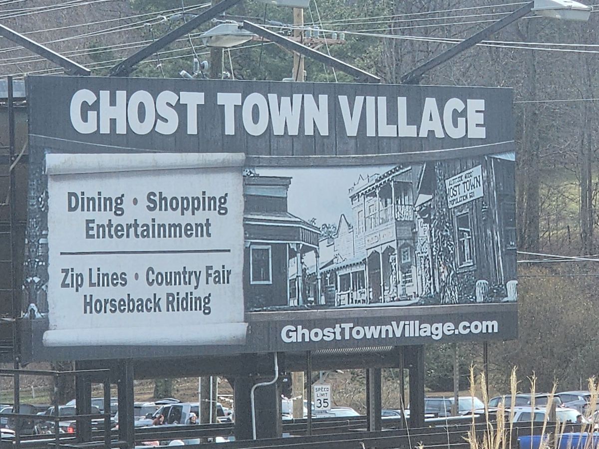 Billboard reading Ghost Town Village, with dining, shopping, entertainment, zip lines, country fair and horseback riding, and an illustration of a Western street.